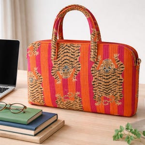Peut inclure: Une sacoche pour ordinateur portable rectangulaire avec un motif à rayures verticales orange et rose vif et un imprimé de tigres répété. Le sac comprend deux poignées en tissu assorties sur le dessus et une fermeture éclair sur le bord supérieur.