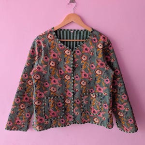 Puede incluir: Una chaqueta acolchada de manga larga con una base verde salvia, rayas verticales verde oscuro y un patrón floral repetitivo en tonos rosa, blanco y amarillo. La chaqueta tiene un cuello redondo, cierre frontal de botones y dos bolsillos.
