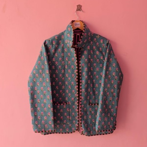 Puede incluir: Una chaqueta acolchada de color azul cerceta apagado con un patrón repetitivo de pequeñas flores rosas con tallos verdes. La chaqueta tiene cuello alto, mangas largas, dos bolsillos delanteros de parche y un borde a rayas en los puños y bordes.