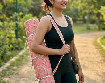 May include: A rolled-up yoga mat featuring a coral-colored fabric with a repeating floral pattern in pink, white, and green. The mat is secured with three matching fabric ties and includes an integrated shoulder strap for carrying.
