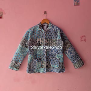 Può includere: Una giacca trapuntata a maniche lunghe con un design patchwork in varie tonalità di blu e bianco con motivi floreali e geometrici. La giacca presenta un colletto alla coreana, una chiusura frontale con bottoni e due tasche. Il testo riporta ShivtextileShop.