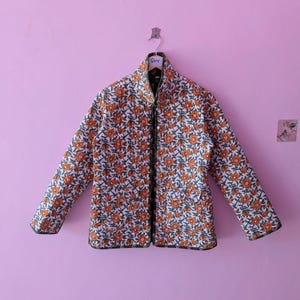 Può includere: Una giacca trapuntata a maniche lunghe con un motivo floreale ripetuto di fiori arancioni e steli verdi su sfondo bianco. La giacca presenta un colletto alto, una chiusura frontale con bottoni e bordi rifiniti in verde scuro.