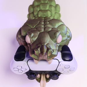 May include: A green and bronze-colored sculpture of a snake head holding a white and black game controller. The snake has a textured, scaly appearance and is mounted on a wall.