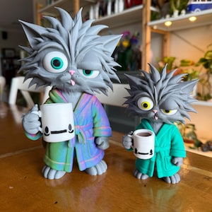 Könnte beinhalten: Zwei graue Katzenfiguren mit struppigem Fell, die jeweils einen Bademantel tragen und eine weiße Tasse halten. Die größere Katze trägt einen bunten, schillernden Bademantel und hat türkisfarbene Augen, die kleinere einen einfarbig türkisen Bademantel.