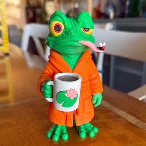 Könnte beinhalten: Eine grüne Froschfigur in einem orangefarbenen Bademantel, die eine weiße Tasse mit einem rosa Lotusblüten- und grünen Seerosenblatt-Motiv hält. Der Frosch hat gelbe Augen mit roten Pupillen, eine lange rosa Zunge und eine kleine Fliege.