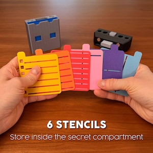 May include: Six colourful plastic stencils in yellow, orange, red, pink, purple, and blue, held in a fan shape. A grey rectangular storage box and a black component with white buttons are visible in the background. Text reads 6 STENCILS Store inside the secret compartment.