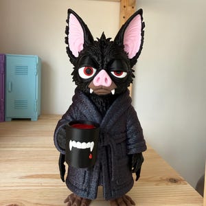 May include: A black bat figurine with large pink-lined ears, red eyes, and a pink snout. The bat wears a dark textured bathrobe and holds a black mug featuring a white graphic of vampire fangs and a single red drop of liquid.