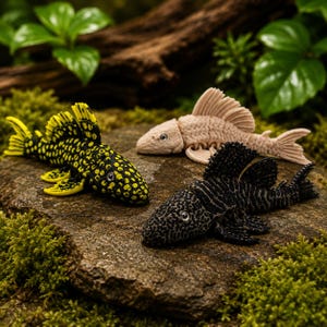 Pode incluir: Três figuras detalhadas de peixes em preto, bege e preto com manchas amarelas, repousando sobre uma superfície rochosa texturizada. Os peixes estão dispostos em um cenário natural com musgo verde e folhagem ao fundo.