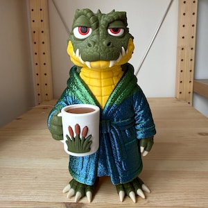 May include: A green crocodile figurine with red eyes and a yellow chest, wearing a metallic blue bathrobe. The crocodile holds a white mug featuring a brown and green cattail plant graphic. The figure has sharp claws and a textured, scaly skin.