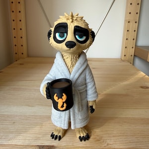 Könnte beinhalten: Eine Erdmännchen-Figur in einem hellgrauen, strukturierten Bademantel, die eine schwarze Tasse mit brauner Flüssigkeit und einem orangefarbenen Skorpion-Motiv auf der Vorderseite hält. Das Erdmännchen hat beiges Fell und blaue Augen.