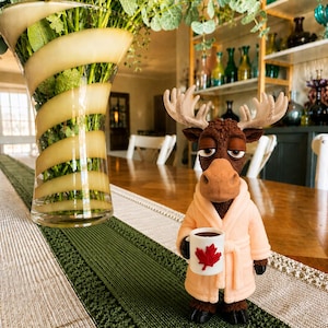 May include: A brown moose figurine with large antlers wearing a light peach-coloured bathrobe. The moose is holding a small white mug featuring a red maple leaf design. The figurine has a sleepy expression with heavy eyelids and dark brown fur.
