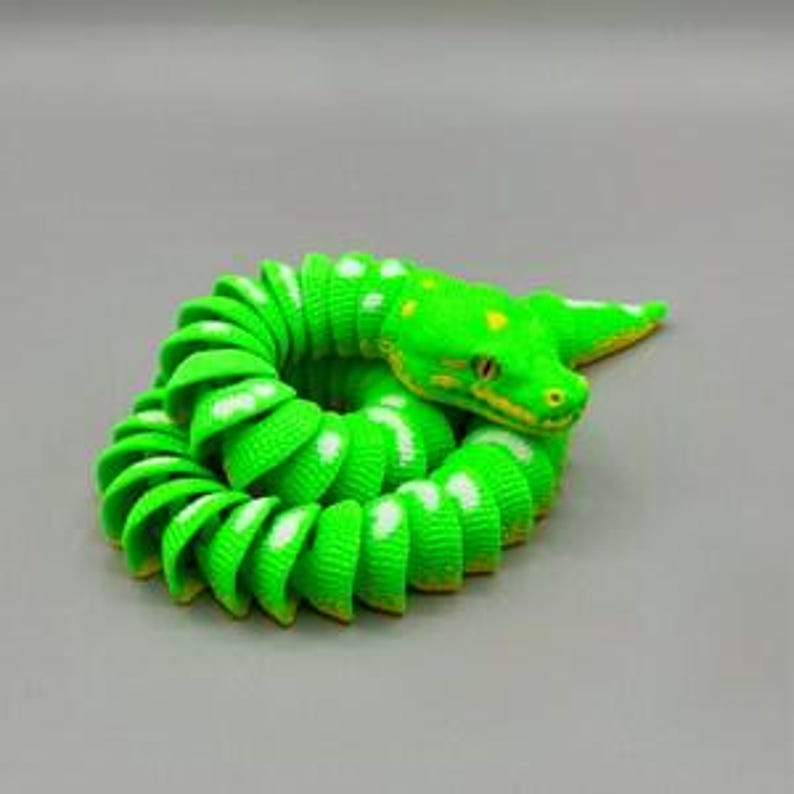 Peut inclure: Figurine de serpent vert enroul&eacute; avec des pois blancs et des d&eacute;tails jaunes. La t&ecirc;te du serpent est relev&eacute;e et ses yeux sont visibles. La figurine est faite d'un mat&eacute;riau c&ocirc;tel&eacute; et repose sur un fond gris.