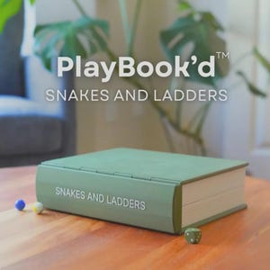 May include: A green board game titled "PlayBook'd Snakes and Ladders" resembling a book. The game is on a wooden table with a die and game pieces. The game's title is printed on the spine and cover.