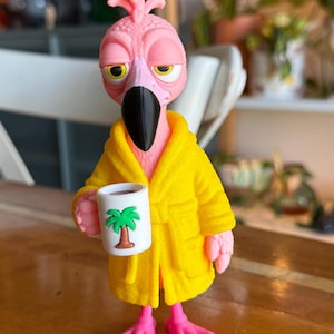 Könnte beinhalten: Eine rosa Flamingo-Figur mit strukturiertem Kopf und schwarzem Schnabel, die einen leuchtend gelben Bademantel trägt. Der Flamingo hält eine kleine weiße Tasse mit einer grünen und braunen Palmen-Grafik, mit müdem Gesichtsausdruck und rosa Füßen.