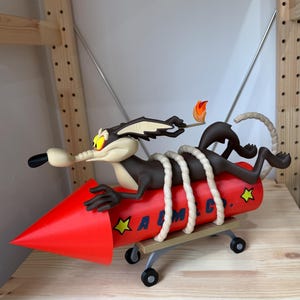 May include: A figurine of a brown coyote with yellow eyes holding a lit match, strapped with white rope to a red rocket labeled ACME CO. with yellow star accents. The rocket is mounted on a small gray four-wheeled cart with a flat platform base.