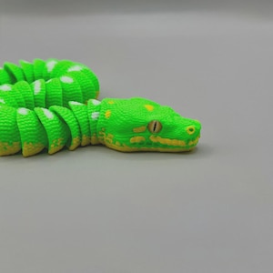 Peut inclure: Un serpent jouet vert vif avec des d&eacute;tails jaunes et des taches blanches. La t&ecirc;te du serpent pr&eacute;sente un &oelig;il r&eacute;aliste et des marques jaunes. Le corps segment&eacute; permet un mouvement flexible, cr&eacute;ant une apparence r&eacute;aliste.