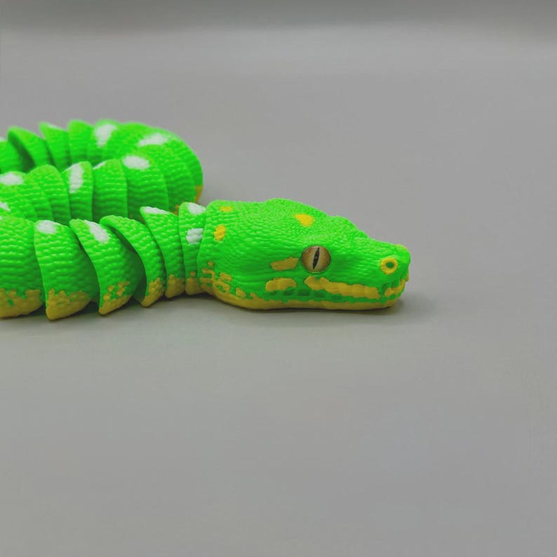Peut inclure: Un serpent jouet vert vif avec des d&eacute;tails jaunes et des taches blanches. La t&ecirc;te du serpent pr&eacute;sente un &oelig;il r&eacute;aliste et des marques jaunes. Le corps segment&eacute; permet un mouvement flexible, cr&eacute;ant une apparence r&eacute;aliste.