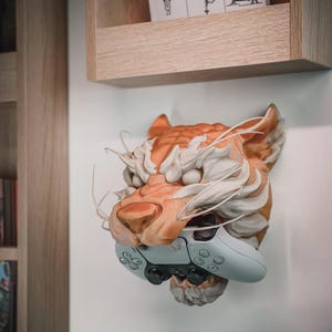 May include: A detailed orange and white tiger head sculpture mounted on a wall, holding a light blue video game controller. The sculpture has intricate fur and facial details. The controller is held in the tiger's mouth.