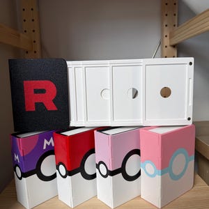 May include: A collection of rectangular storage cases featuring designs inspired by spherical monster capture devices. One black case displays a red letter R, while others feature purple, red, pink, and light blue colour schemes with circular patterns.
