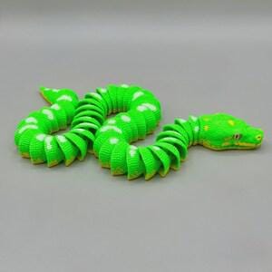 Peut inclure: Un serpent jouet vert et blanc avec un corps segment&eacute; et un dessous jaune. Le serpent a des taches blanches et une t&ecirc;te d&eacute;taill&eacute;e avec des accents jaunes. Le jouet est pos&eacute; sur une surface grise.