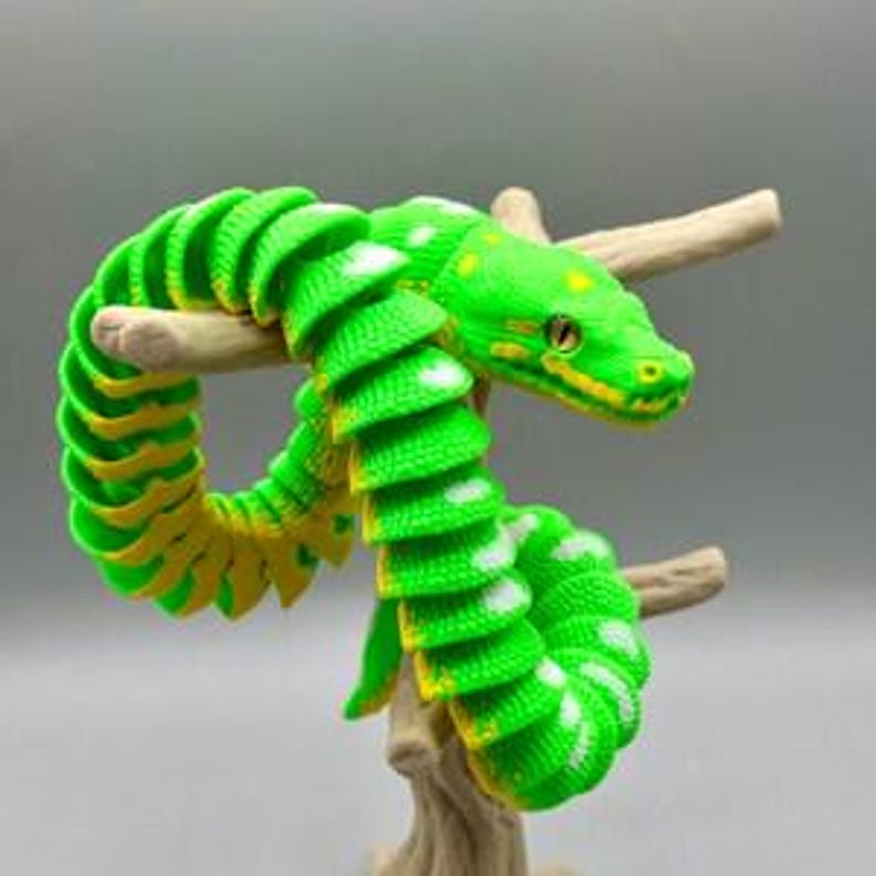 Peut inclure: Un jouet serpent articul&eacute; vert vif et jaune enroul&eacute; autour d'une petite branche marron clair. Le serpent pr&eacute;sente des taches blanches et des accents jaunes. La t&ecirc;te du serpent est d&eacute;taill&eacute;e avec des yeux jaunes et une langue fourchue. Le fond est de couleur neutre.