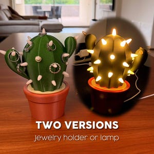 May include: Two cactus-shaped decorative items. One is a green jewelry holder with rings on its arms, the other is a lamp with warm white lights. Both are in terracotta-coloured pots. Text reads "TWO VERSIONS jewelry holder or lamp."