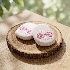 Op de afbeelding: Twee ovale, roze en wit gestreepte compactpoeders met de woorden "WITH GOD" en "anything is possible" in roze letters. De compactpoeders liggen op een houten schijf met een natuurlijke rand, op een houten oppervlak.