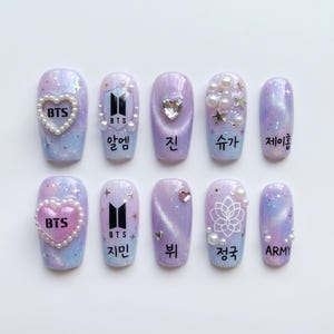 May include: A set of ten artificial nails with a light purple and blue galaxy design. Each nail features embellishments like pearls, hearts, stars, and the BTS logo. Some nails have Korean text.