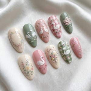 Könnte beinhalten: Ein Set aus zehn oval geformten Kunstnägeln, die jeweils mit einem einzigartigen Design bemalt sind. Die Designs umfassen Schwäne, Blumenarrangements und Vichy-Muster in Pastelltönen wie Rosa, Grün und Creme. Die Nägel liegen auf weißem Satin.
