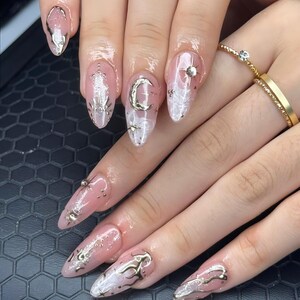 May include: Almond-shaped nails featuring a sheer pink base with white marbled accents, metallic silver crescent moon and star charms, and small silver studs. The design includes raised silver liquid-like detailing across the nail surfaces.
