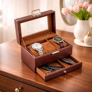 May include: A brown leather watch box with a clear glass top, open to reveal two watches on brown velvet cushions. A drawer below holds watch accessories. The box is on a wooden surface.