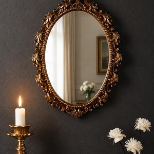 Può includere: Uno specchio da parete ovale con una cornice dorata decorata, caratterizzata da intricati intagli a forma di foglia e un bordo interno perlato. Lo specchio riflette una stanza con una finestra, tende e un vaso di fiori bianchi.