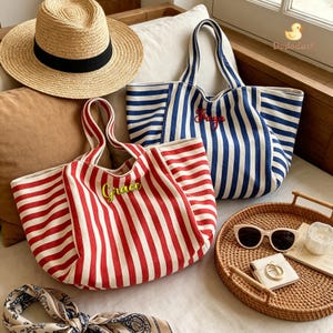 May include: A straw hat with a black band, a red and white striped canvas tote bag with the name Grace embroidered in green, and a blue and white striped canvas tote bag with the name Freya embroidered in red, alongside sunglasses and a scarf.