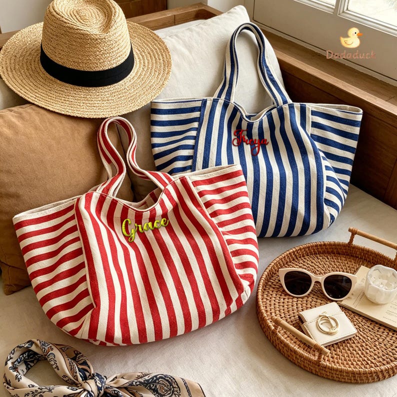 May include: A straw hat with a black band, a red and white striped canvas tote bag with the name Grace embroidered in green, and a blue and white striped canvas tote bag with the name Freya embroidered in red, alongside sunglasses and a scarf.