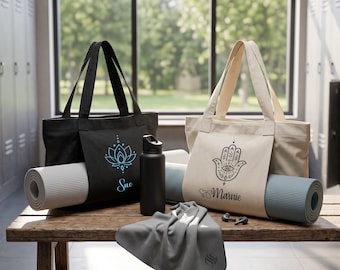 May include: A black tote bag with a blue lotus graphic and the name Sue, and a beige tote bag with a black hamsa hand graphic and the name Marnie. Both bags hold rolled yoga mats. A black water bottle and a gray towel are also present.