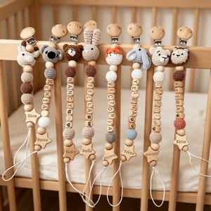 May include: A selection of personalised baby dummy clips. Each clip has a wooden bead chain, a crocheted animal head, and a wooden star with a name. Animals include a giraffe, koala, bear, bunny, fox, elephant, lion, and a crocheted ball.