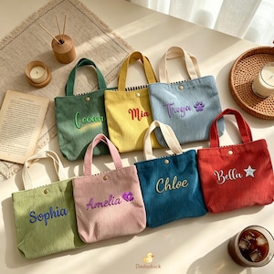 May include: A selection of corduroy tote bags in different colours, each embroidered with a name. The bags have contrasting handles and a button closure. Colours include green, yellow, blue, pink, and red. The bags are personalised with names like "Sophia", "Mia", and "Bella".