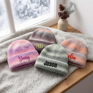 May include: Four knit beanies with folded cuffs, each featuring embroidered names. One is pink and purple with 'Daisy', one is brown and grey with 'Ali', one is grey with 'Jason', and one is orange and pink with 'Amelia' embroidered.