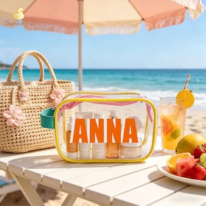 May include: A clear cosmetic bag with the name "ANNA" in orange letters, filled with skincare products. A woven tote bag with pink flower accents sits beside it on a white table. A refreshing drink and fruit plate are also on the table.
