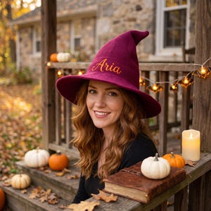 May include: A deep burgundy witch hat featuring a textured waffle-knit fabric and the name Aria embroidered in a gold script font on the front. The hat has a tall, pointed crown and a wide, slightly curved brim, designed for a costume accessory.