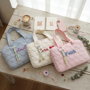 May include: Three rectangular quilted laptop bags with heart-shaped stitching patterns in light blue, cream, and light pink. Each bag features a personalized embroidered name, Ameli, Cora, or Faith, and a colorful beaded tassel zipper pull.