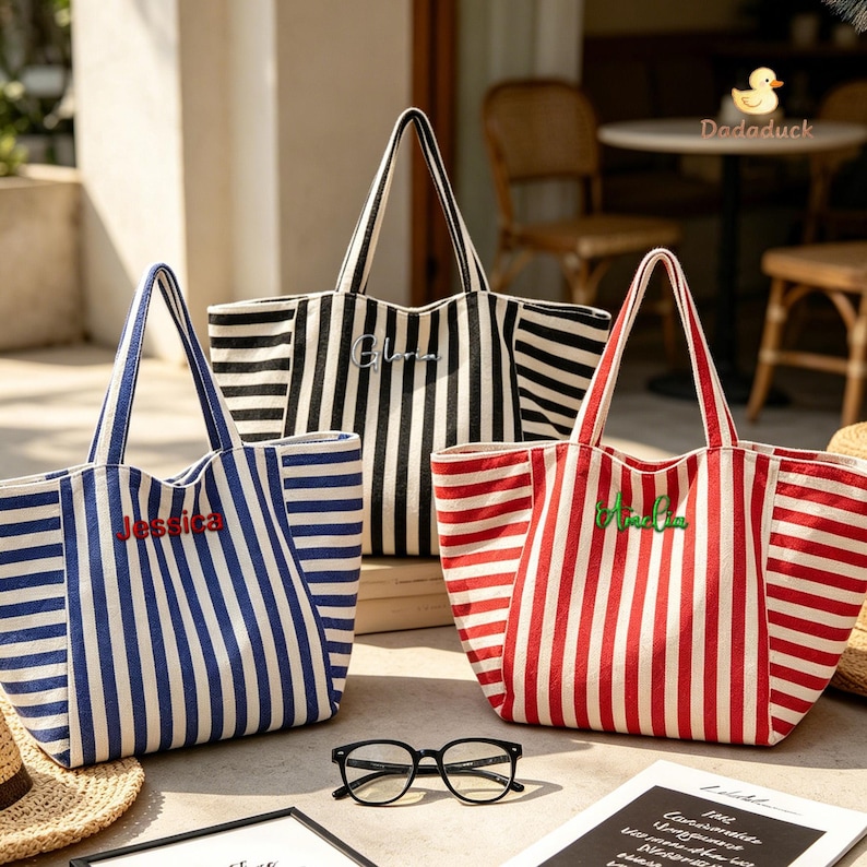 May include: Three canvas tote bags with vertical stripes in blue, black, and red. Each bag features two handles and a name embroidered on the front: Jessica in red, Gloria in white, and Aveline in green. Black-rimmed eyeglasses rest in the foreground.