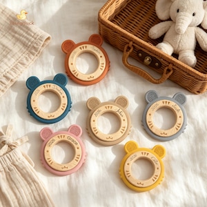 Op de afbeelding: Een verzameling kleurrijke bijtringen voor baby's in verschillende tinten, waaronder blauw, oranje, roze, beige, grijs en geel. Elke bijtring heeft een berenvormig ontwerp met een houten middenstuk en de naam van een kind. De bijtringen liggen op een witte ondergrond.