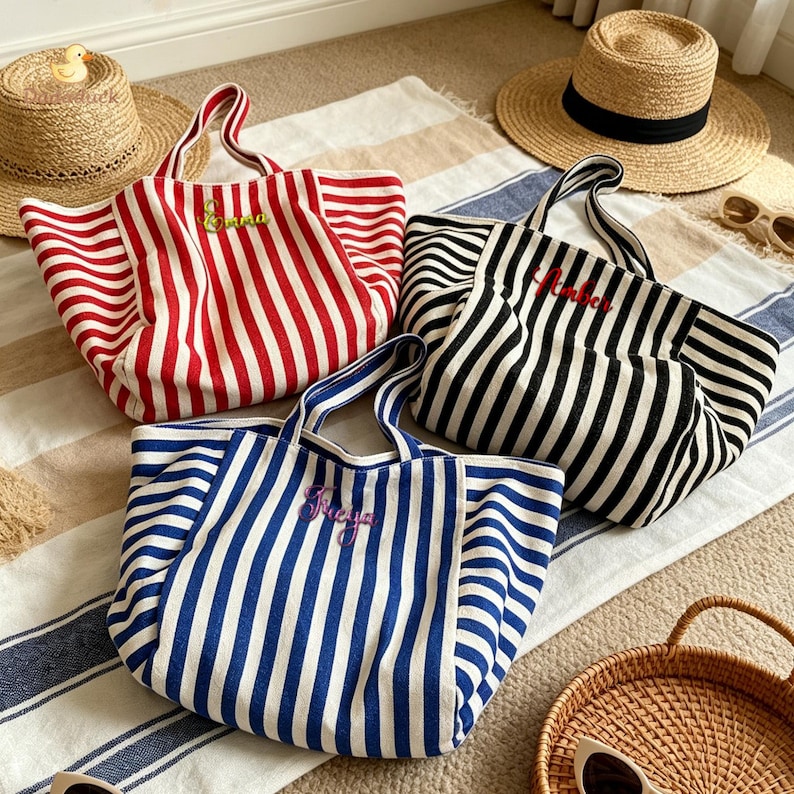 May include: Three canvas tote bags with vertical stripes in red, black, and blue. Each bag features two handles and a name embroidered on the front: Emma in green, Amber in red, and Freya in purple. The bags are arranged on a striped fabric surface.