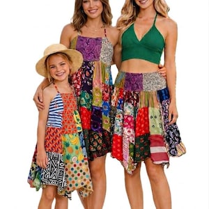 May include: Three women and a child wearing patchwork dresses and a green crop top. The dresses feature a variety of colorful patterns and prints. The child wears a straw hat. The dresses are knee-length.