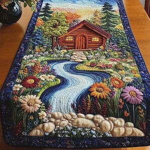 Könnte beinhalten: Ein rechteckiger Tischläufer aus Stoff mit einer detaillierten, gesteppten Landschaftsszene. Das Design zeigt eine braune Blockhütte, einen geschwungenen blauen Fluss, einen Steinpfad und bunte Blumen, eingefasst von einem dunkelblauen Rand.