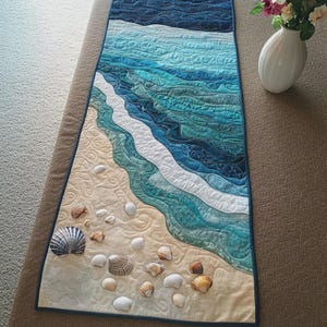 Peut inclure: Un chemin de table rectangulaire en tissu présentant une scène océanique matelassée avec des vagues bleu et sarcelle s'écrasant sur un rivage beige sablonneux. La plage est ornée de divers coquillages en tissu en relief sur la surface.