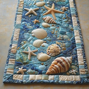 May include: A rectangular fabric table runner featuring a quilted ocean-themed design with various appliqued seashells and starfish in shades of white, tan, and brown against a textured blue background with intricate wave-like stitching patterns.