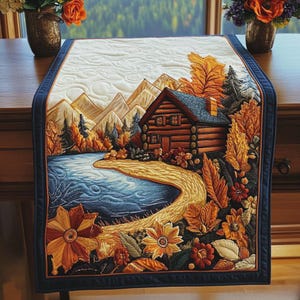 May include: A rectangular fabric table runner featuring a quilted landscape scene of a log cabin beside a blue lake, surrounded by autumn foliage, mountains, and trees in shades of orange, yellow, and brown, finished with a dark blue border.