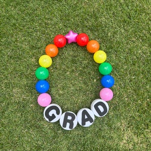 Könnte beinhalten: Eine farbenfrohe Abschlussdekoration, kreisförmig auf grünem Gras angeordnet. Sie besteht aus einem rosa Stern, roten, orangefarbenen, gelben, grünen, blauen und rosa Kugeln sowie dem Wort "GRAD" auf weißen Kreisen.